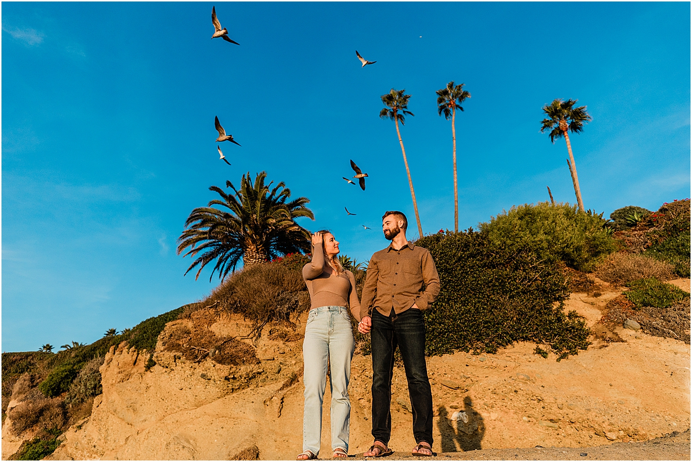 Laguna beach couples photography at Treasure Island Park. Couple holding hands on rocks in front of palm trees and seagulls flying.