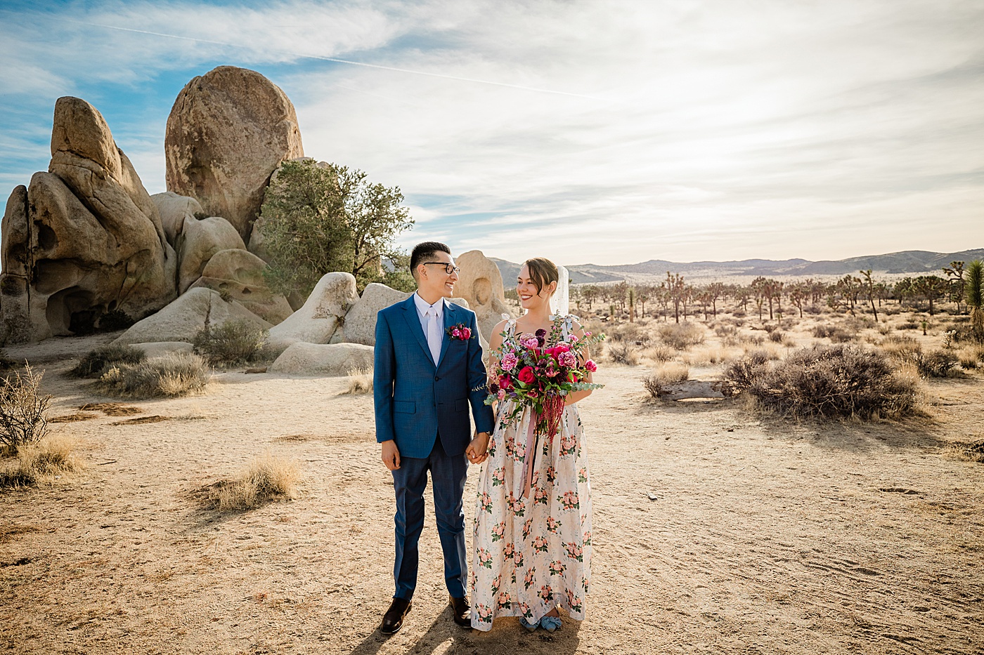 Joshua Tree elopement. Groom in a blue suit, and bride in a floral wedding dress hold hands laughing together in front of a rock formation.
