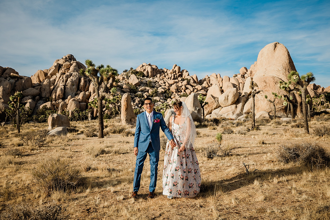 Joshua Tree elopement. A groom in a blue suit, and bride in a floral wedding dress hold hands laughing together in front of a rock formation.