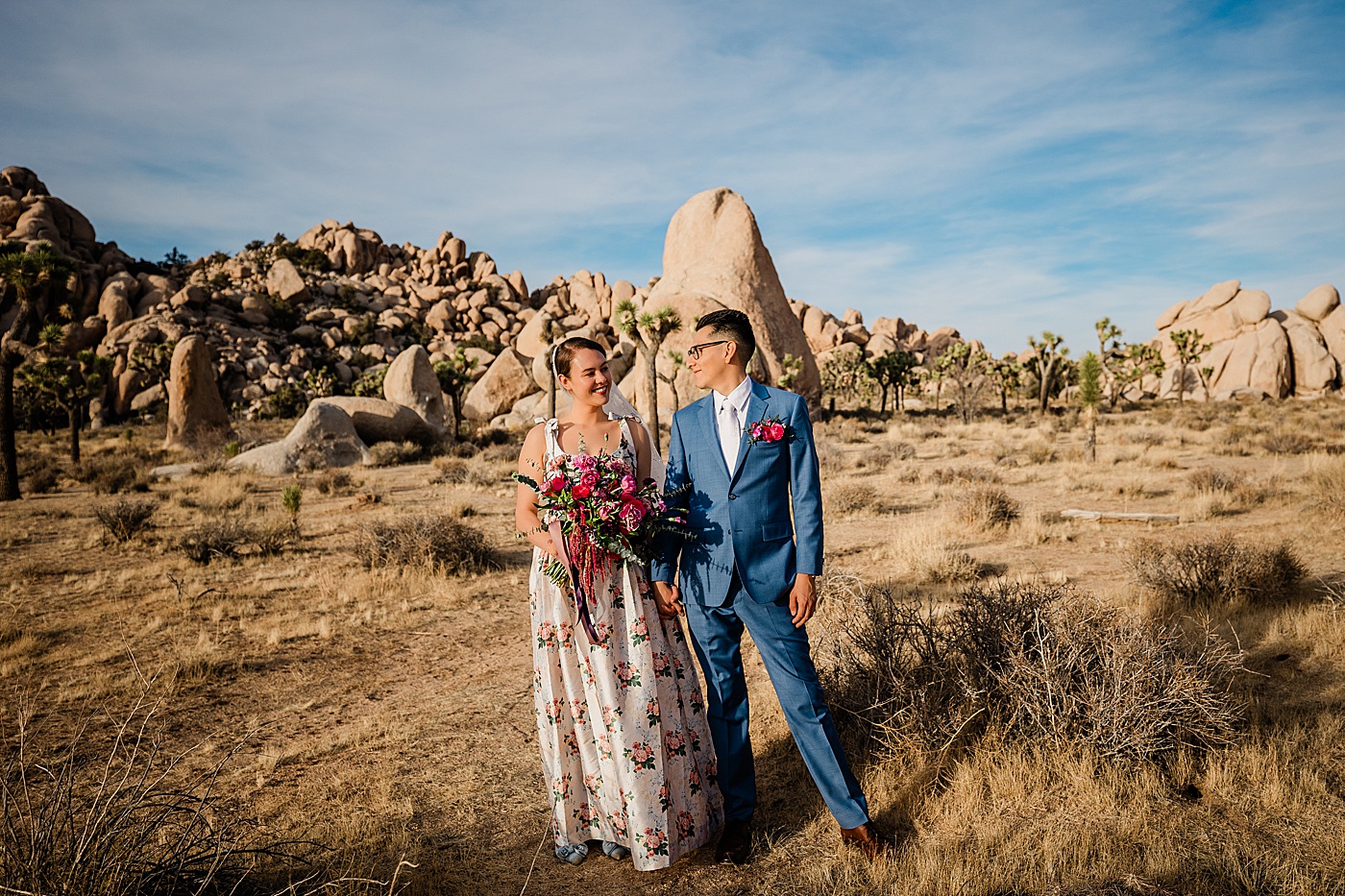 Joshua Tree elopement. A groom in a blue suit, and bride in a floral wedding dress hold hands laughing together in front of a rock formation.