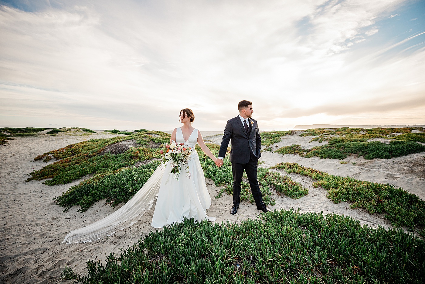 Coronado beach elopement at the sand dunes. A bride and groom holding hands and looking away from each other.